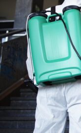 Unrecognizable person in white chemical protection suit with reservoir disinfecting public corridor to stop spreading highly contagious corona virus.