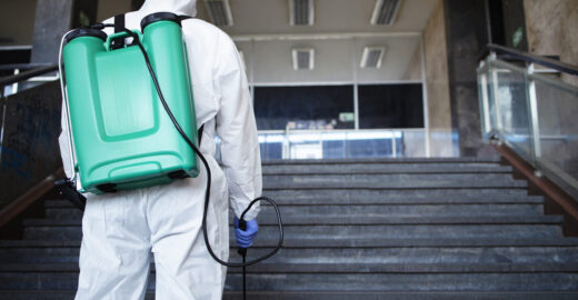Unrecognizable person in white chemical protection suit with reservoir disinfecting public corridor to stop spreading highly contagious corona virus.